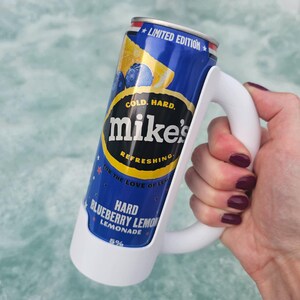 May include: A can of Mike's Hard Blueberry Lemonade in a white mug-style holder. The can is blue with a yellow lemon and blueberry graphic. Text on the can reads "Cold. Hard. Refreshing." and "Hard Blueberry Lemon Lemonade."