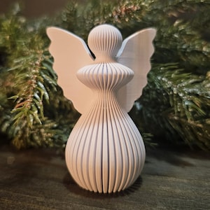 May include: A white, 3D-printed angel figurine with a stylized design. The angel has a rounded head, a flared body, and large, flat wings. The figurine is made of vertical, parallel lines, giving it a textured appearance. The background features green pine branches.