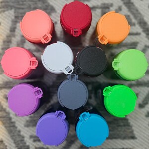 May include: A collection of colorful, round, portable speakers. The speakers are in various colors, including pink, red, orange, white, black, green, purple, blue, and gray. Each speaker has a circular top with a small handle and a latch.
