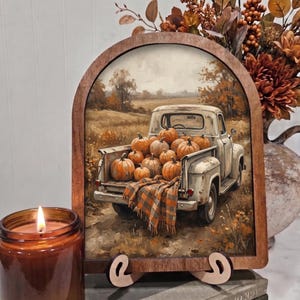 May include: An arched wooden frame containing a print of a vintage white pickup truck filled with orange pumpkins in a field. A plaid blanket hangs over the tailgate. The scene features autumn colors, trees, and a soft, painterly texture throughout.