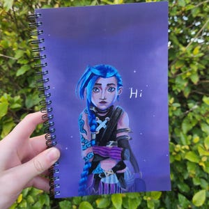 May include: A spiral-bound notebook with a character illustration. The character has blue hair and is set against a purple background with the word "Hi". The character wears a black outfit with purple accents. The notebook is held against green foliage.