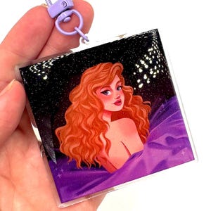 May include: Square acrylic keychain featuring a woman with red hair, set against a starry night sky and purple fabric. The keychain has a purple clasp. The artwork has a glittery effect.