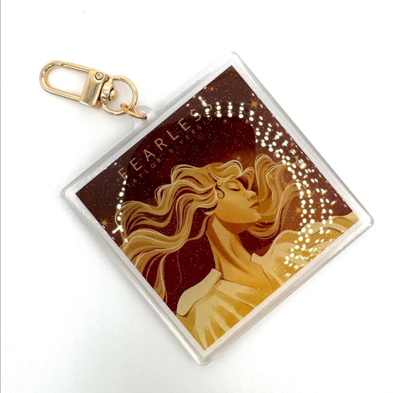 May include: A clear, square keychain with a gold clasp. The keychain features a graphic of a woman with flowing blonde hair and the text "FEARLESS TAYLOR'S VERSION" in white. The background is a dark brown with gold glitter.