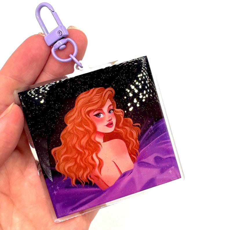 May include: Square acrylic keychain featuring a woman with red hair, set against a starry night sky and purple fabric. The keychain has a purple clasp. The artwork has a glittery effect.
