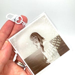 May include: A clear acrylic keychain featuring a sepia-toned illustration of a woman in a white dress. The keychain has a silver-colored clasp and a small metal ring. The artwork depicts a woman looking over her shoulder.