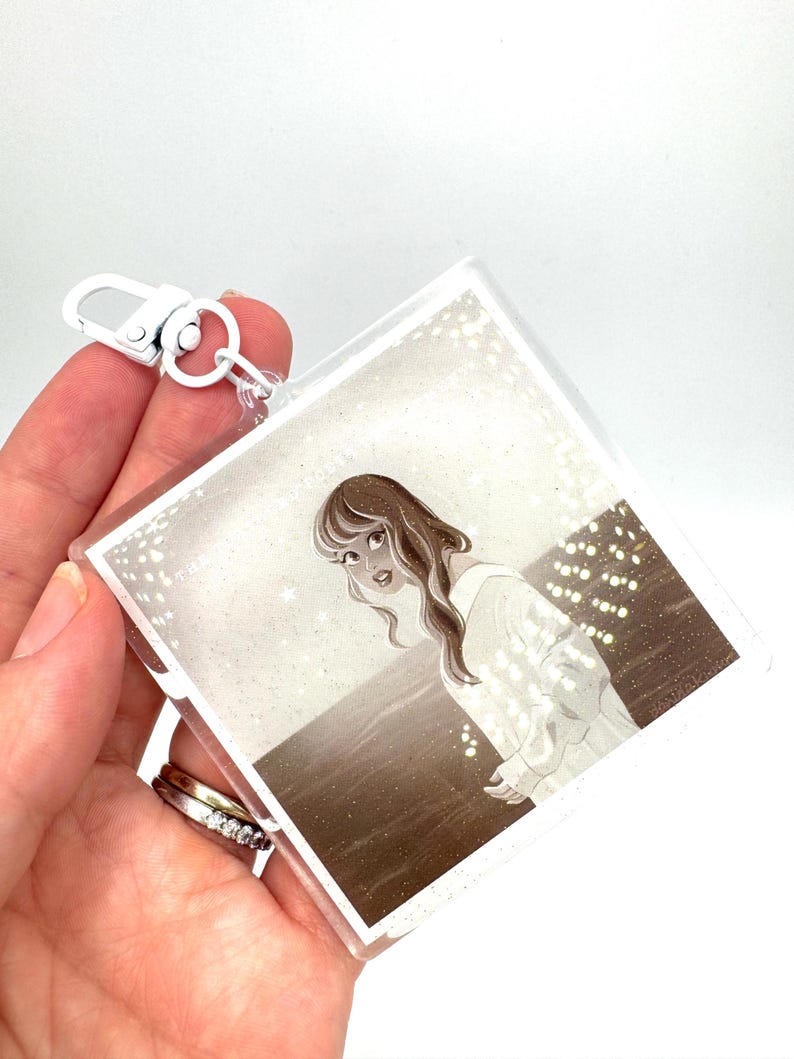 May include: A clear acrylic keychain featuring a sepia-toned illustration of a woman in a white dress. The keychain has a silver-colored clasp and a small metal ring. The artwork depicts a woman looking over her shoulder.