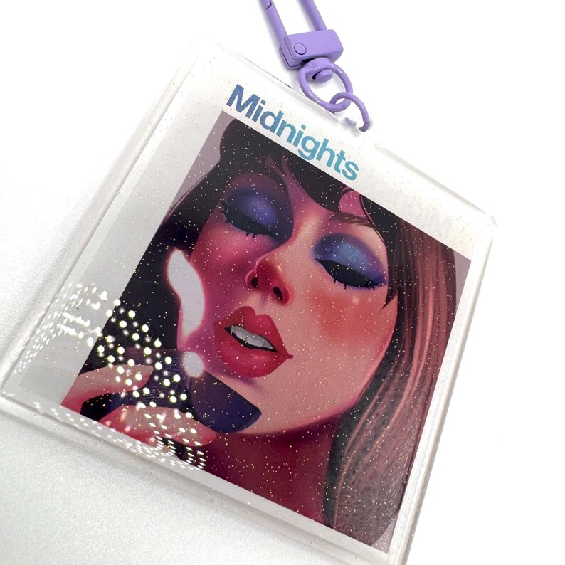 May include: A clear, glittery keychain with a portrait of a woman with blue eyeshadow and red lipstick. The word "Midnights" is printed in light blue. The keychain has a purple clasp.