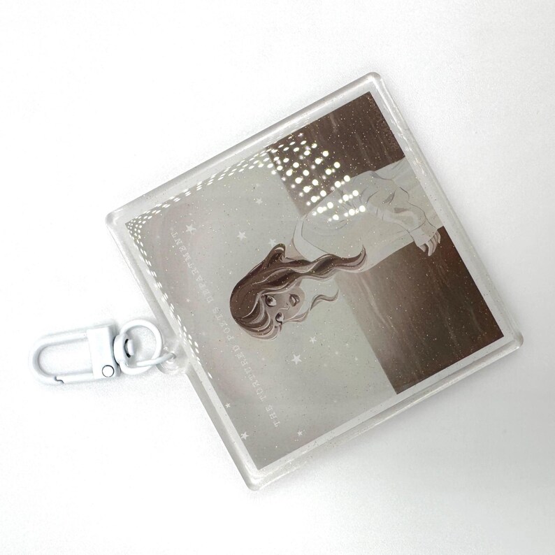 May include: A clear acrylic keychain with a silver clasp. The square pendant features a sepia-toned illustration of a woman with long hair, with the text "THE TORMENTED POETS DEPARTMENT" around the border.