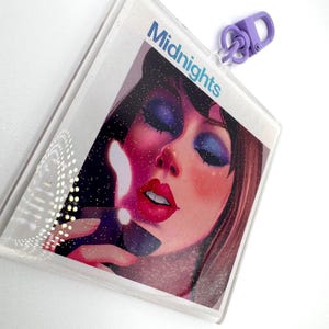 May include: A clear, glittery keychain featuring a portrait of a woman with blue eyeshadow and red lipstick. The word "Midnights" is printed in blue. A purple clasp is attached to the top.