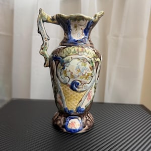 Puede incluir: Jarra decorativa de cerámica con asa. La jarra presenta un diseño floral con detalles en azul, amarillo y rosa. La base es marrón y el borde está festoneado. La jarra mide aproximadamente 20 cm de altura.