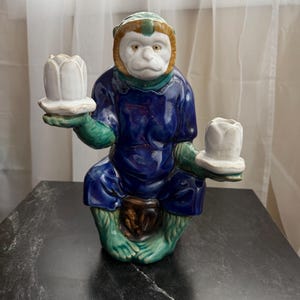 May include: A ceramic monkey figurine with a white face, green and blue clothing, and two white candle holders. The monkey is seated and holding the candle holders. The figurine is on a black surface.