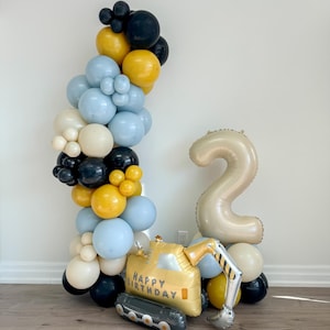 May include: A balloon arch with blue, yellow, and black balloons. A large gold number 2 balloon and a yellow construction vehicle balloon with the text "Happy Birthday" are in the foreground.