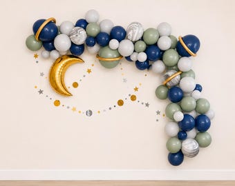 May include: A space-themed balloon arch featuring a large gold crescent moon foil balloon, navy blue, sage green, white, and marble-patterned balloons, and several balloons with gold rings to resemble planets, accented by small gold and silver star cutouts.