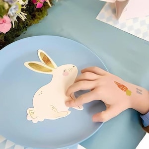 May include: A blue plate with a white bunny cutout on top. The bunny has gold ears and a pink nose. A child's hand is reaching for the bunny.