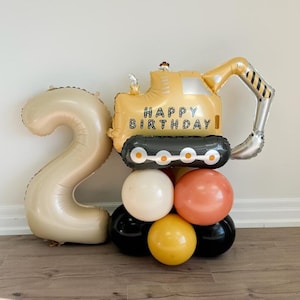 May include: A gold number two balloon with a yellow construction vehicle balloon on top. The vehicle balloon has the words "Happy Birthday" on it. The balloons are surrounded by smaller balloons in black, white, yellow, and orange.