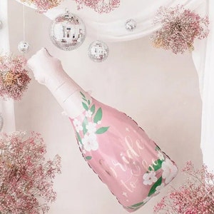 May include: Pink champagne bottle shaped balloon with white flowers and green leaves. The balloon has the text "Bride to Be" on it. The balloon is hanging from a white ceiling with silver disco balls and pink flowers.