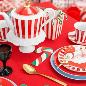May include: A red and white striped teapot with a gold lid and handle, a white saucer, a red and white striped candy cane holder, a red and white striped teacup and saucer, a gold spoon, and two candy cane place card holders on a red tablecloth.
