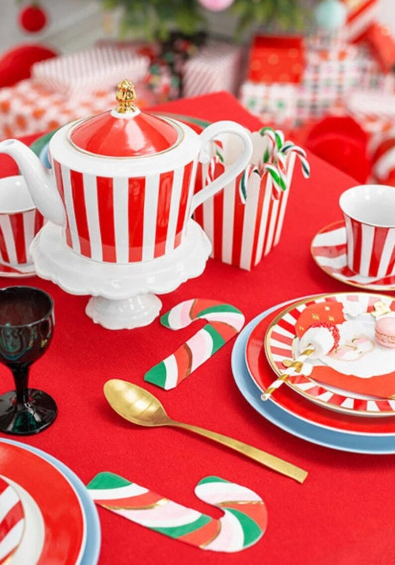 May include: A red and white striped teapot with a gold lid and handle, a white saucer, a red and white striped candy cane holder, a red and white striped teacup and saucer, a gold spoon, and two candy cane place card holders on a red tablecloth.