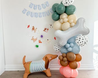 Puede incluir: Un arco de globos con un globo en forma de hueso azul y blanco, globos verdes, crema y marrones, y un globo blanco con huellas de patas negras. Un globo de dachshund marrón y blanco está en primer plano. La pancarta dice "Feliz cumpleaños".
