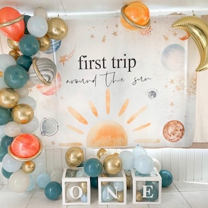 Puede incluir: Una decoración de fiesta de primer cumpleaños con un arco de globos azul, blanco y naranja, un telón de fondo con el texto "primer viaje alrededor del sol" y tres cajas blancas con las letras "ONE" en dorado.