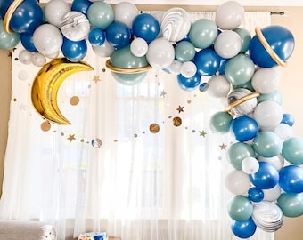 Puede incluir: Un arco de globos hecho de globos azules, blancos y grises con detalles dorados. Los globos están dispuestos en forma de media luna y están decorados con estrellas doradas. El arco está colgado frente a una ventana con cortinas blancas.