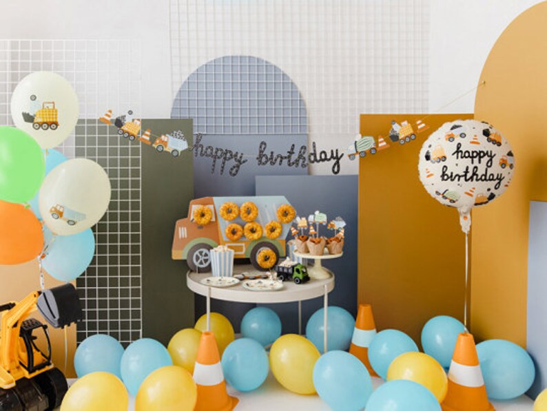 K&ouml;nnte beinhalten: Eine festliche Geburtstagsfeier mit Luftballons, einem Spielzeugbagger und einem Tisch mit Donuts und Cupcakes. Die Szene umfasst ein Banner mit der Aufschrift "Happy Birthday" und einen Ballon mit der gleichen Aufschrift. Das Farbschema umfasst Gelb, Blau und Orange.