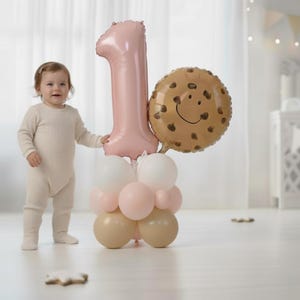 May include: A festive arrangement of balloons for a first birthday celebration. The display includes a large pink number one balloon, a cookie-shaped balloon with a smiling face, and a cluster of smaller balloons in white, pink, and beige. A baby in a beige onesie stands nearby.