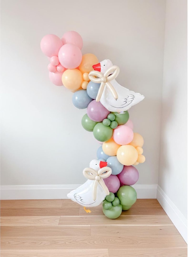 May include: A whimsical balloon garland featuring a variety of pastel-colored balloons in pink, yellow, green, and blue. Two white goose-shaped balloons with red beaks and cream-colored bows are incorporated into the design, creating a charming party decoration.