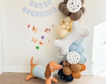 May include: A dog-themed birthday party setup featuring a dachshund balloon, a balloon arch with paw print balloons, and a banner that says "Happy Birthday". The color scheme includes brown, blue, and white.