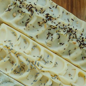 May include: Four bars of handmade soap, each with a different design. The top bar has a swirl design with dried herbs and flowers. The second bar has a swirl design with lavender buds. The third bar has a swirl design with poppy seeds. The bottom bar has a swirl design with a white base.