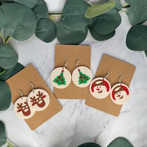 Puede incluir: Tres pares de pendientes con temática navideña. El primer par presenta un árbol de Navidad verde con adornos rojos. El segundo par presenta un muñeco de nieve con una bufanda roja y ojos negros. El tercer par presenta un reno con una nariz roja y cuernos marrones.