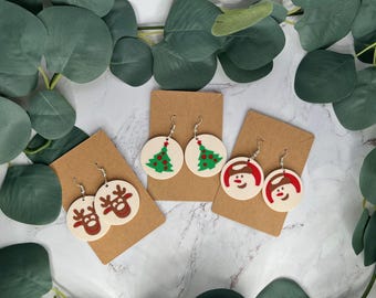 May include: Three pairs of Christmas-themed earrings. The first pair features a green Christmas tree with red ornaments. The second pair features a snowman with a red scarf and black eyes. The third pair features a reindeer with a red nose and brown antlers.