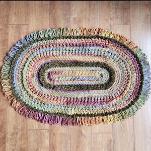 May include: Oval-shaped, handmade crochet rug with a fringed edge. The rug features a blend of colors, including green, brown, purple, orange, and yellow. The rug is placed on a wooden floor.