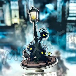 May include: A black and white acrylic charm featuring a street lamp with three small black figures with yellow eyes. The charm is on a clear acrylic base.