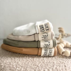 Könnte beinhalten: Stapel aus vier gerippten Stoff-Haarbinden in Weiß, Beige, Olivgrün und Braun. Jede Haarbinde hat ein weißes Stoff-Etikett mit schwarzem Text. Die Etiketten lauten "Trouble Maker", "Hej Hej", "Hej Hej Echt" und "Jetzt?"