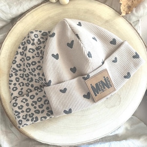 Könnte beinhalten: Zwei beige Baby-Mützen. Eine Mütze hat ein Leopardenmuster, die andere ein Herzmuster. Die Mütze mit Herzmuster hat ein braunes Lederetikett mit dem Wort "MINI". Die Mützen liegen auf einer Holzoberfläche.