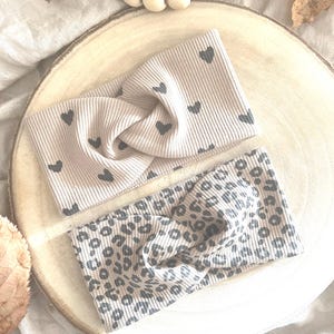 Könnte beinhalten: Zwei beige gerippte Stirnbänder. Eines hat ein Muster aus kleinen grauen Herzen, das andere einen grauen Leoparden-Print. Die Stirnbänder sind in der Mitte verdreht und bilden so ein stilvolles Accessoire. Sie bestehen aus weichem Rippstoff.