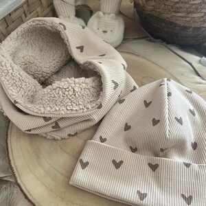 Pode incluir: Um conjunto de gorro e gola de malha canelada bege. O gorro e a gola s&atilde;o decorados com pequenos cora&ccedil;&otilde;es castanhos. A gola tem um interior macio e fofo. O conjunto &eacute; exibido em uma superf&iacute;cie de madeira.
