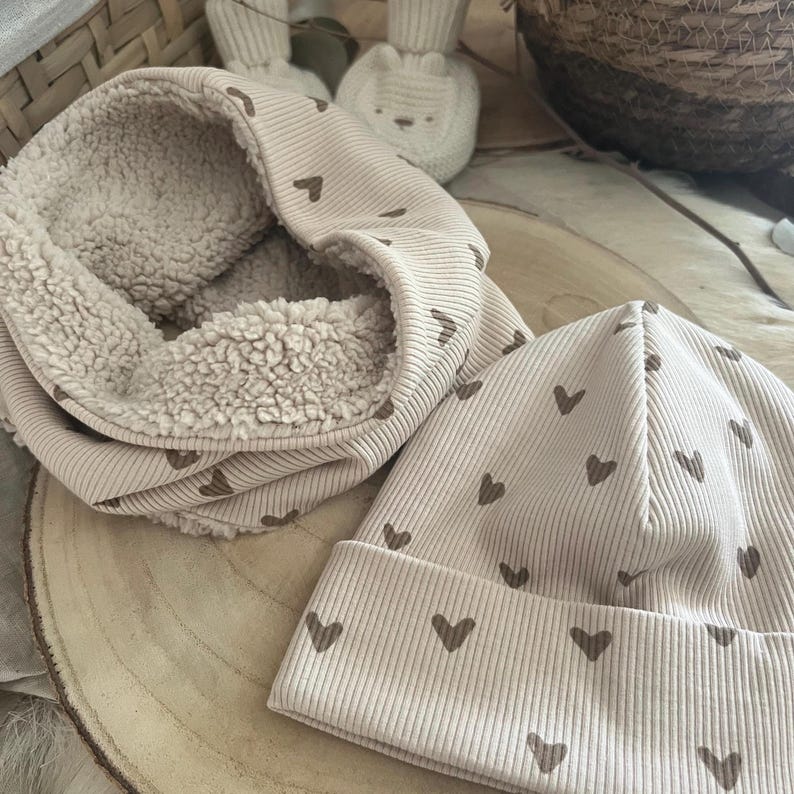 Pode incluir: Um conjunto de gorro e gola de malha canelada bege. O gorro e a gola s&atilde;o decorados com pequenos cora&ccedil;&otilde;es castanhos. A gola tem um interior macio e fofo. O conjunto &eacute; exibido em uma superf&iacute;cie de madeira.