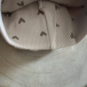 Pode incluir: Um gorro de malha canelada bege com uma aba branca. O interior do gorro apresenta um padr&atilde;o de pequenos cora&ccedil;&otilde;es castanhos. O gorro est&aacute; sobre uma superf&iacute;cie de madeira clara e texturizada.