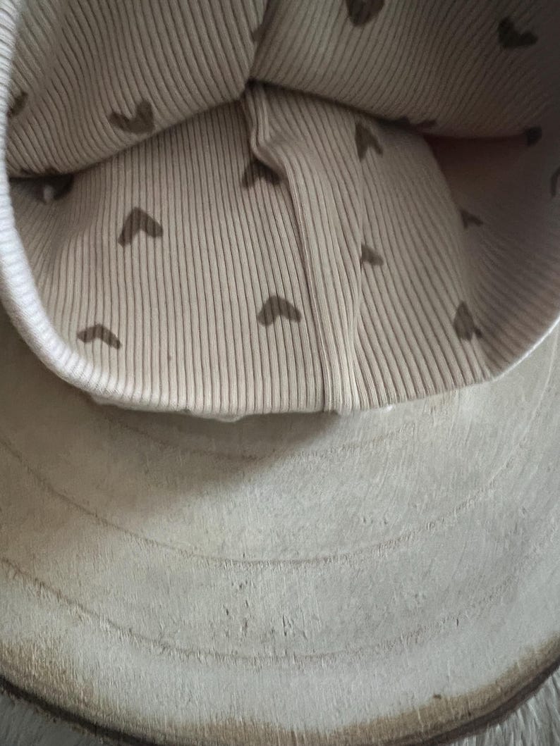 Pode incluir: Um gorro de malha canelada bege com uma aba branca. O interior do gorro apresenta um padr&atilde;o de pequenos cora&ccedil;&otilde;es castanhos. O gorro est&aacute; sobre uma superf&iacute;cie de madeira clara e texturizada.