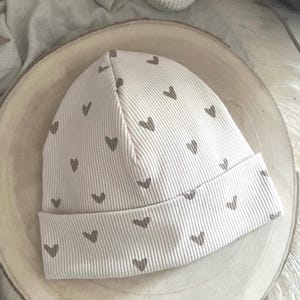 Pode incluir: Um gorro de malha canelada creme com uma aba dobrada. O chap&eacute;u &eacute; estampado com pequenos cora&ccedil;&otilde;es cinzentos espa&ccedil;ados uniformemente. O chap&eacute;u foi concebido para aquecer e proporcionar conforto.
