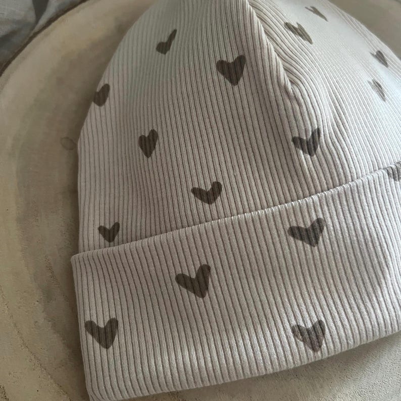 Pode incluir: Um gorro de malha canelada bege claro com uma aba dobrada. O gorro &eacute; decorado com pequenos padr&otilde;es em forma de cora&ccedil;&atilde;o castanho escuro. O gorro &eacute; feito de um material macio e el&aacute;stico, adequado para beb&eacute;s e crian&ccedil;as.