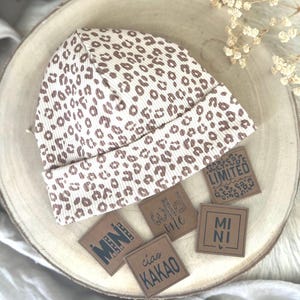 Könnte beinhalten: Eine beige-braune Beanie-Mütze mit Leopardenmuster und gerippter Textur. Mehrere kleine braune Lederetiketten mit schwarzem Text sind um die Mütze angeordnet. Die Etiketten lauten "MINI", "Wild One", "LIMITED", "ciao KAKAO" und "MI NI".