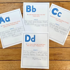 Puede incluir: Cuatro tarjetas blancas con bordes azules y letras azules grandes A, B, C y D. Cada tarjeta tiene un versículo bíblico y un ejercicio de trazado. Las tarjetas están sobre una superficie de madera.