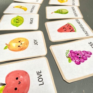 Pode incluir: Um conjunto de cartas coloridas com ilustra&ccedil;&otilde;es de frutas e palavras como "amor", "alegria", "paz", "paci&ecirc;ncia", "bondade", "fidelidade", "mansid&atilde;o" e "autocontrole".