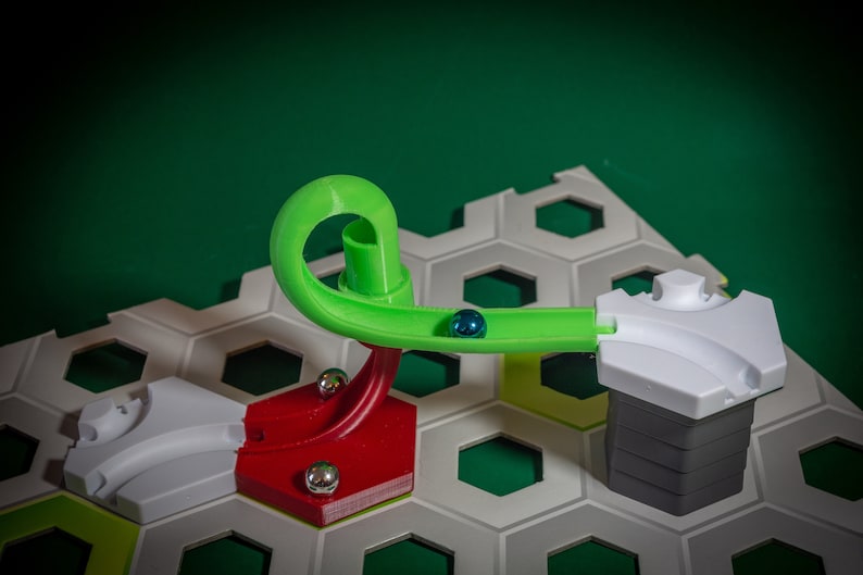 Puede incluir: Una pieza de pista de m&aacute;rmol verde curva con una canica azul rodando a trav&eacute;s de ella. La pista est&aacute; unida a una base roja y blanca, y la base se coloca en un tablero de juego hexagonal gris.