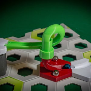 Puede incluir: Una pieza de pista de pl&aacute;stico verde con un bucle para un juego de carrera de canicas. La pieza de pista est&aacute; conectada a una base roja con una pieza de pista blanca. Hay tres canicas plateadas y una canica azul en la base roja.