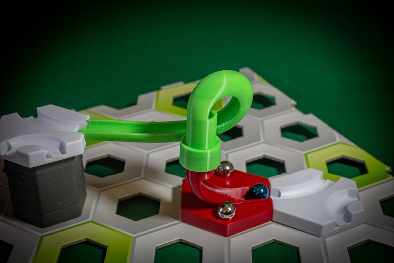 Puede incluir: Una pieza de pista de pl&aacute;stico verde con un bucle para un juego de carrera de canicas. La pieza de pista est&aacute; conectada a una base roja con una pieza de pista blanca. Hay tres canicas plateadas y una canica azul en la base roja.