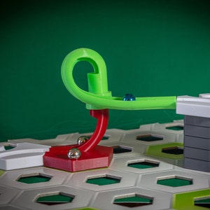 Puede incluir: Una pista de m&aacute;rmol verde y roja con una torre blanca y gris. La pista est&aacute; hecha de pl&aacute;stico y tiene un bucle. La torre est&aacute; hecha de bloques apilados. La pista est&aacute; colocada sobre un tablero hexagonal blanco y verde.